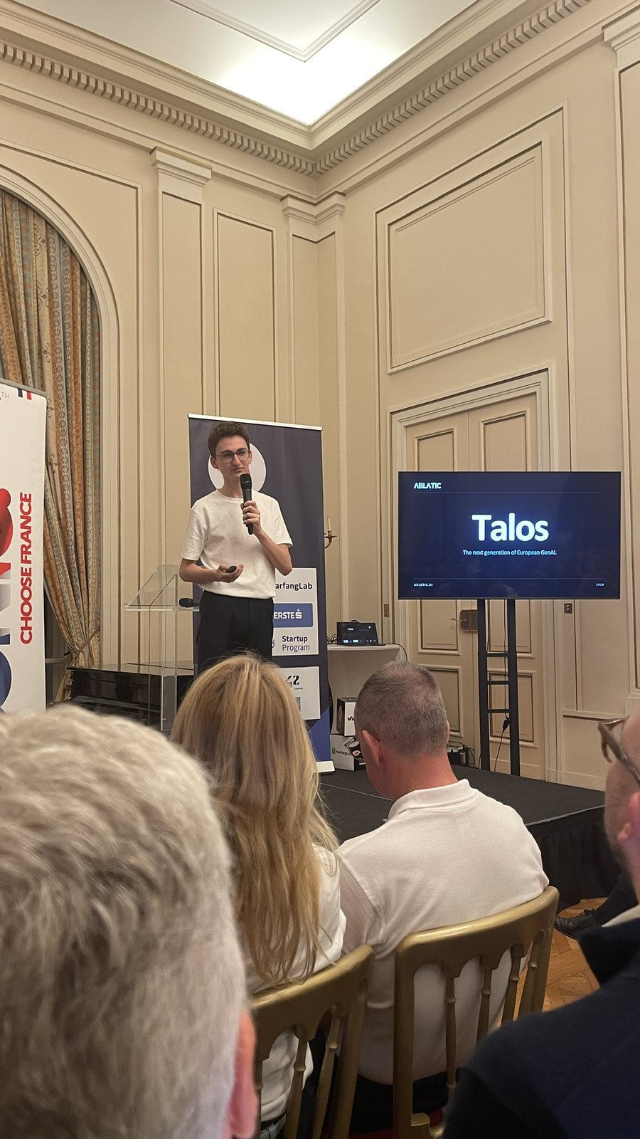 An Ablatic co-founder speaks into a microphone next to a screen showing the slide Talos, the next generation of European GenAI.