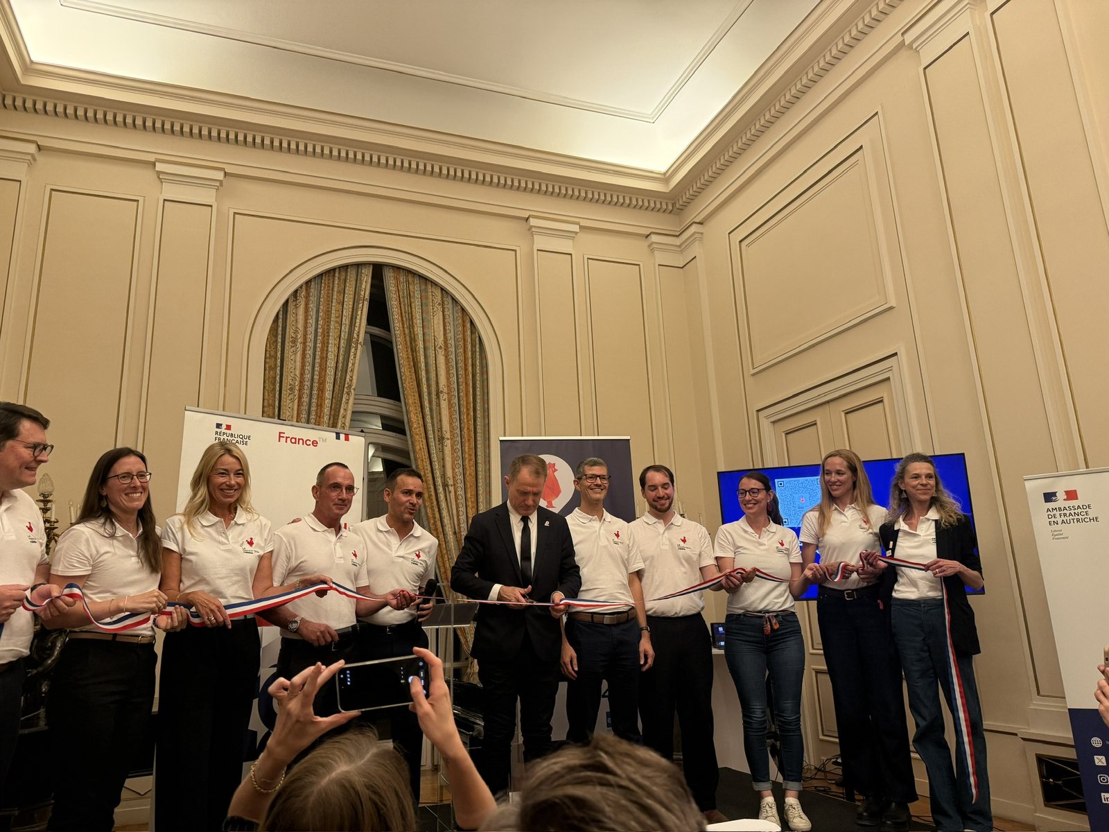 French Ambassador Matthieu Peyraud cuts a blue, white and red ribbon held by members of la French Tech Vienna in white polo shirts, in a cream-panelled room at the French Embassy.